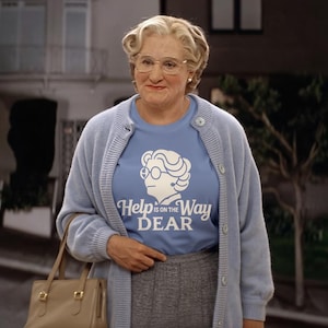 May include: A person wearing a light blue cardigan over a light blue t-shirt with the text "Help is on the Way Dear" and a graphic of a woman's face. The person is also wearing glasses and a gray skirt, and carrying a tan handbag.