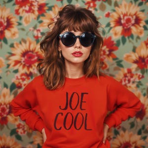 May include: A woman wearing a red sweatshirt with the text "JOE COOL" in black lettering. She is wearing round black sunglasses and has brown hair with bangs.