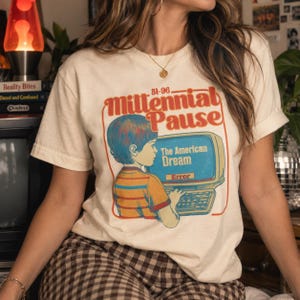 May include: A cream-colored short-sleeve t-shirt featuring a graphic print of a child at a computer. The text reads 81-96 Millennial Pause, The American Dream, and Error. The illustration uses orange, blue, and yellow tones on the shirt fabric.