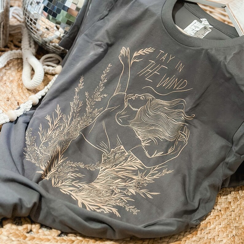 May include: A charcoal gray crew neck t-shirt featuring a light tan line art graphic of a person with flowing hair amidst tall grasses. The text TAY IN THE WIND is printed in a stylized font above the figure, with a visible size tag at the neck.