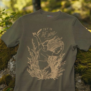 May include: An olive green short-sleeved t-shirt featuring a light-colored line art graphic of a person with flowing hair standing among tall grasses. The text "TAY IN THE WIND" is printed above the figure in a stylized, cursive-inspired font.