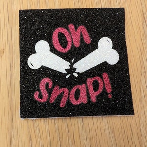 May include: A black glitter patch with a white broken bone design and the text "Oh Snap!" in pink glitter.