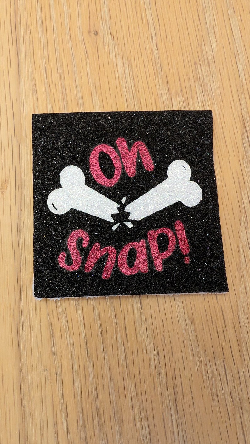 May include: A black glitter patch with a white broken bone design and the text "Oh Snap!" in pink glitter.