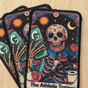 May include: Rectangular patch featuring a colorful sugar skull design with a skeleton holding a green sports drink. The text "The Athletic Trainer" is at the bottom. The background is black with stars, moons, and flowers.