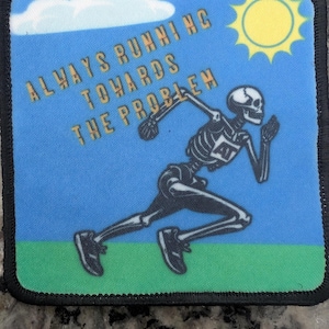 Puede incluir: Parche cuadrado con fondo azul que presenta un gráfico de un esqueleto corriendo. El texto "ALWAYS RUNNING TOWARDS THE PROBLEM" está en letras doradas. Un sol amarillo y una nube blanca están en la esquina superior derecha. El parche tiene un borde negro.