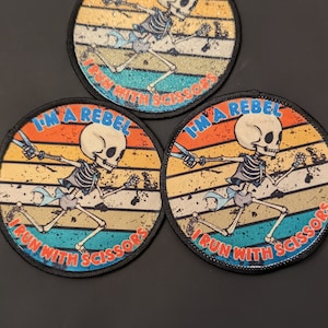 Puede incluir: Tres parches redondos para planchar con un esqueleto corriendo con tijeras. Cada parche tiene un fondo de puesta de sol retro con el texto "I'm a Rebel I Run With Scissors".