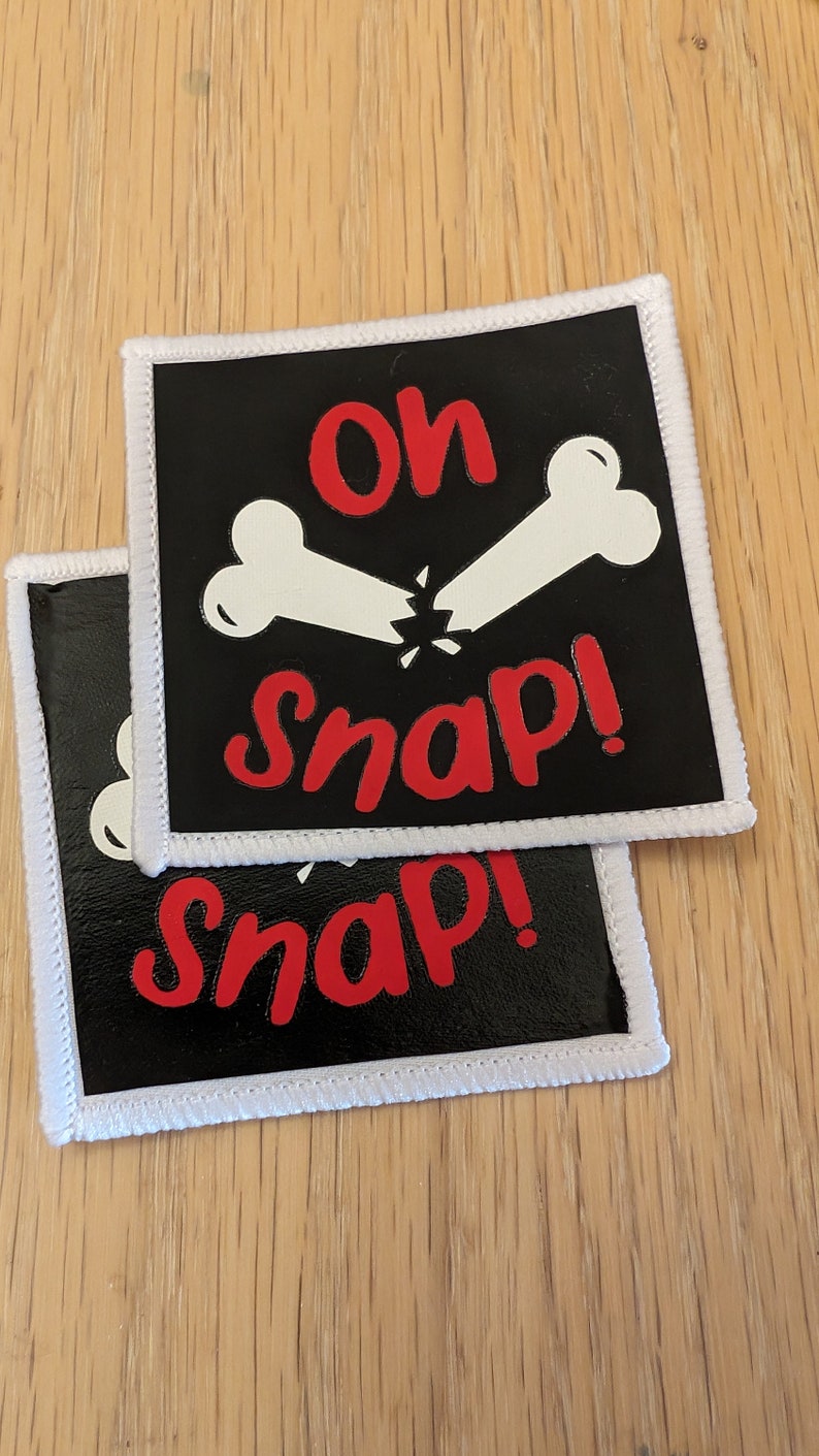 May include: Two black fabric patches with white stitching around the edges. The patches have a white bone design with a red "Oh Snap!" text on the top patch and a red "Snap!" text on the bottom patch.