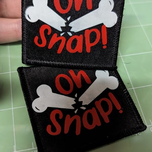 May include: Two black fabric patches with white bone designs and red text that reads "Oh Snap!"