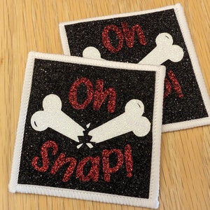 May include: Two black and white iron-on patches with white bone designs and red glitter text that reads "Oh Snap!"