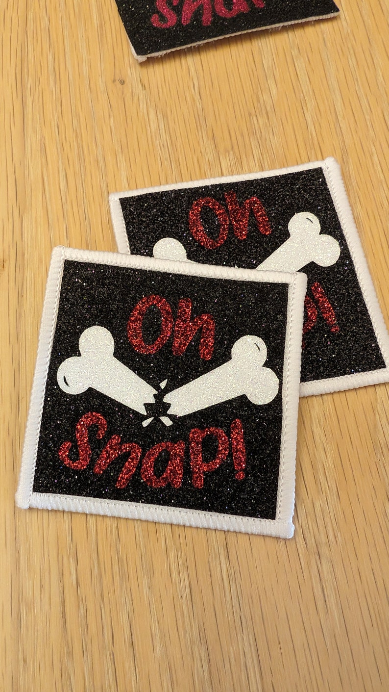 May include: Two black and white iron-on patches with white bone designs and red glitter text that reads "Oh Snap!"