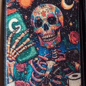 May include: A black and white patch with a colorful sugar skull holding a green can with the letter "G" on it. The skull is surrounded by roses and stars. The text "The Athletic Trainer" is at the bottom of the patch.