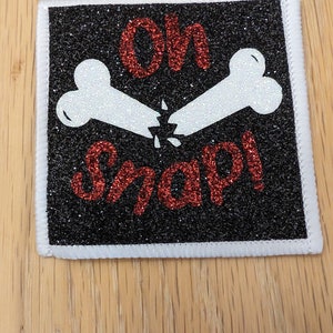 May include: An iron-on patch with a glittery finish. The patch is black and white with two white bones crossed over each other and the text "Oh Snap!" in red glitter.