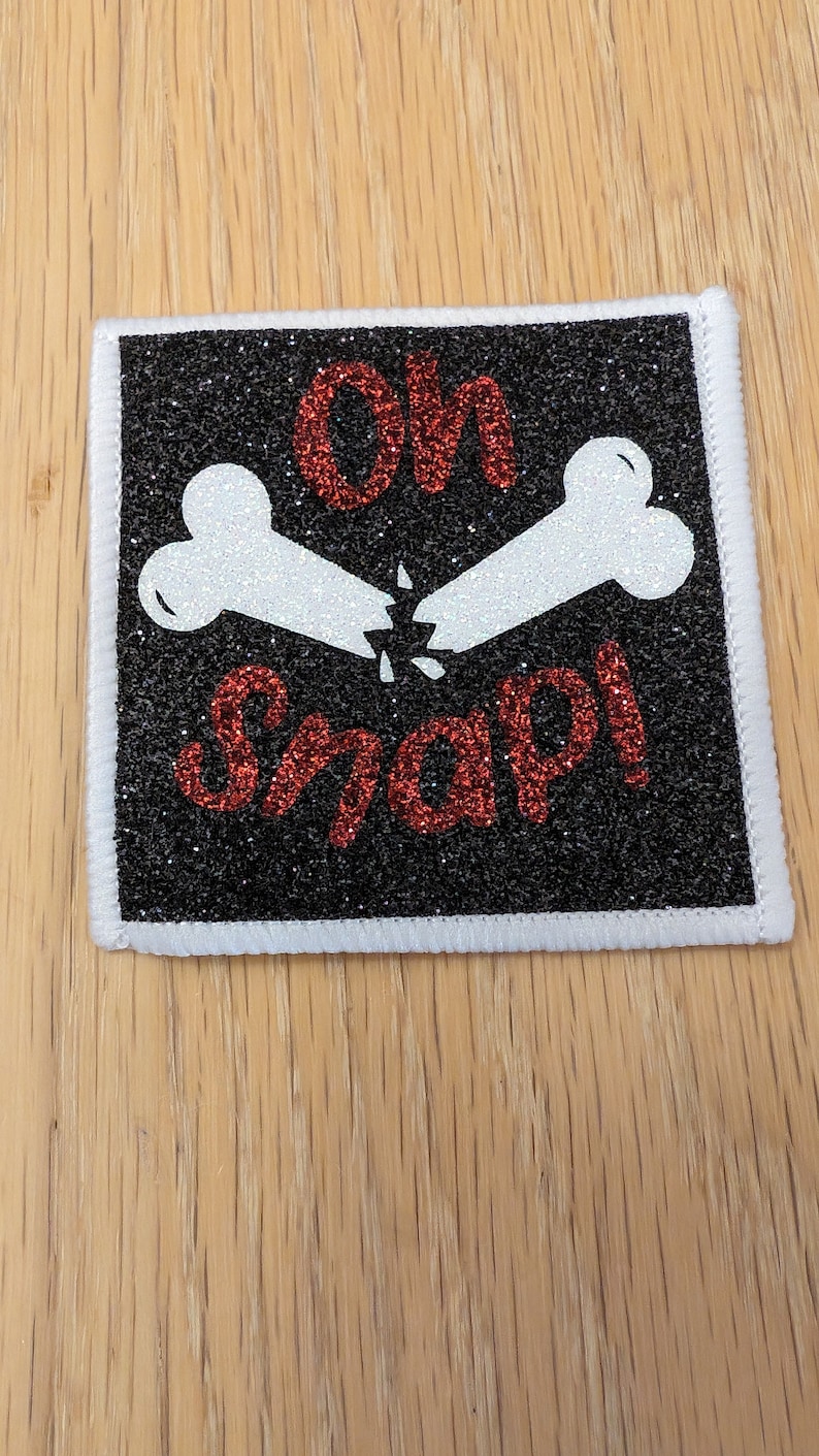 May include: An iron-on patch with a glittery finish. The patch is black and white with two white bones crossed over each other and the text "Oh Snap!" in red glitter.