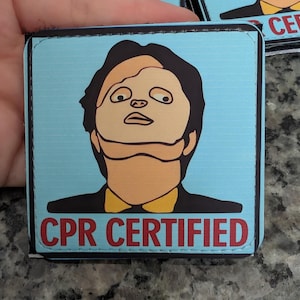 May include: Square, light blue item with a cartoon image of a person in a black suit and yellow shirt. The text "CPR CERTIFIED" is in red letters at the bottom. Several similar items are visible.