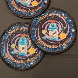 May include: Three round black and purple patches with a white, blue, and orange cartoon image of a Gatorade bottle with a face and the text "Mercury is in Gatorade Again".