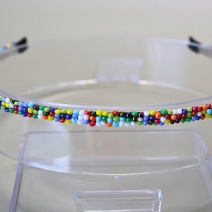 May include: A colorful beaded headband. The headband is made of a clear plastic band with a row of small, multi-colored beads. The beads are in shades of red, blue, green, yellow, and white. The headband is displayed on a clear stand.