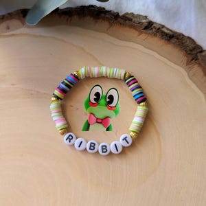 May include: A colorful beaded bracelet with the word "RIBBIT" spelled out in white letter beads. The bracelet features a cartoon frog charm and is accented with gold-colored beads.