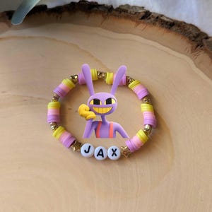 Puede incluir: Una colorida pulsera de cuentas con un personaje de conejo de dibujos animados morado con guantes amarillos y una amplia sonrisa. La pulsera incluye cuentas amarillas, rosas y doradas, con el nombre "JAX" escrito con cuentas de letras blancas. Cuentas en forma de estrella.