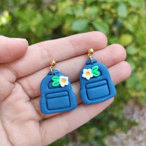 Puede incluir: Un par de pendientes colgantes con forma de mochilas azules en miniatura. Cada mochila presenta una pequeña flor blanca con centro amarillo y dos hojas verdes en la parte superior. Los pendientes cuelgan de postes metálicos dorados.