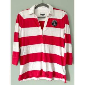 May include: A long-sleeve polo shirt featuring horizontal red and white stripes, a white collar, and a partial button placket. A circular black and white patch with the text Abercrombie & Fitch New York is located on the upper left chest area.