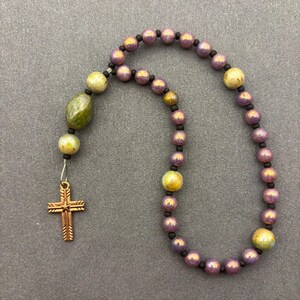 May include: A beaded necklace featuring a series of iridescent purple and yellow spherical beads separated by small black spacers. One end of the strand includes a larger, irregular green stone bead and a metallic copper-toned cross pendant.