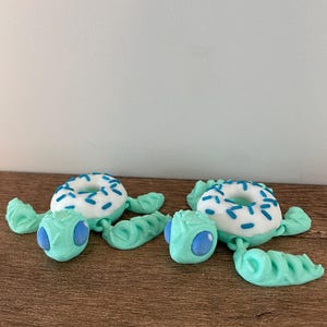 May include: Two sea turtle figurines with light mint green bodies and large blue eyes. Each turtle features a white doughnut-shaped shell on its back, decorated with small blue sprinkles. The figures have articulated flippers and heads.