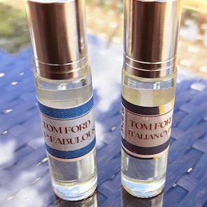 May include: Two clear glass bottles with silver caps and labels. The labels are white with black text and a blue and white design. The text on the labels reads "TOM FORD" and "ITALIAN CYPRESS" and "TOM FORD" and "FABULOUS".