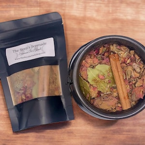 May include: A black pouch with the text "The Siren's Serenade" and "Summon the Wild" on the front. The pouch contains dried herbs and a cinnamon stick. The pouch is sitting on a wooden surface next to a black cauldron filled with dried herbs and a cinnamon stick.