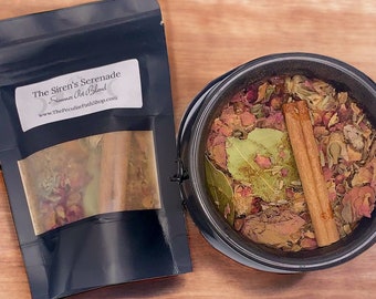 May include: A black pouch with the text "The Siren's Serenade" and "Summon the Wild" on the front. The pouch contains dried herbs and a cinnamon stick. The pouch is sitting on a wooden surface next to a black cauldron filled with dried herbs and a cinnamon stick.