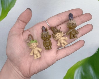May include: Four small troll dolls with varying skin tones and crystal accents on their heads. Each troll has a unique hairstyle made of moss and small twigs. The dolls are held in a hand, with a green leafy background.