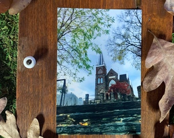 May include: A framed photograph of a church building with a steeple, surrounded by fall foliage. The church is made of stone and has a pointed roof. The leaves are brown and orange.