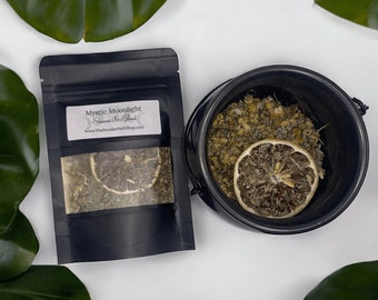 May include: A black pouch with the text "Mystic Moonlight" and "Summer Solstice Blend" on the front. The pouch contains dried herbs and a dried lemon slice. The pouch is sitting on a white surface next to a black cauldron filled with dried herbs and a dried lemon slice.