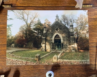 May include: A vintage wooden box with a hinged lid, containing a photograph of a stone building with arched windows and a door. The box is surrounded by brown leaves.