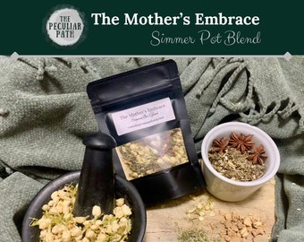May include: A close-up of a white bowl filled with dried herbs and star anise, a black mortar and pestle filled with dried white flowers, and a black pouch with a white label that reads "The Mother's Embrace Simmer Pot Blend".