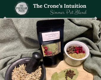 May include: A black pouch with a white label that reads "The Crone's Intuition Simmer Pot Blend" and a website address. The pouch is on a wooden surface with a mortar and pestle, a small white bowl with red berries and black peppercorns, and a bay leaf with cloves. The background is a green fabric.