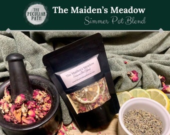 May include: A black pouch with a white label that reads "The Maiden's Meadow Simmer Pot Blend" and a website address. The pouch contains dried flowers, herbs, and citrus fruit. The pouch is sitting on a wooden surface with a mortar and pestle, a bowl of lavender, and a green fabric.