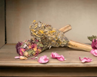 May include: Two bundles of dried herbs and flowers tied together with a wooden stick. The bundles are made of dried lavender, rose petals, and other herbs. The bundles are on a wooden surface with pink flower petals scattered around them.