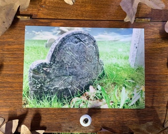 May include: A close-up photograph of a weathered gray headstone with a heart carved into it. The headstone is surrounded by green grass and is set against a backdrop of a blue sky with white clouds.