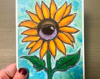May include: A colorful illustration of a sunflower with an eye in the center. The petals are orange, the eye is purple and brown, and the background is blue. The flower is on a white card.