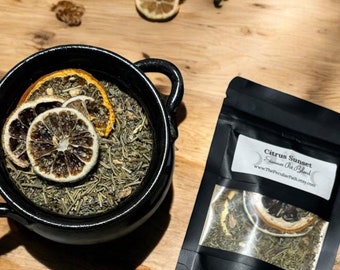 May include: A black ceramic bowl filled with green tea leaves and dried citrus slices. A black pouch with the text "Citrus Sunset" and a clear window showing tea and citrus is next to the bowl. The items are on a wooden surface.
