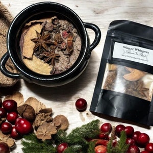 May include: A black ceramic bowl filled with spices and star anise, alongside a black pouch labeled "Winter Whispers". The scene includes scattered cranberries, pine sprigs, and other natural elements, creating a festive, seasonal arrangement.