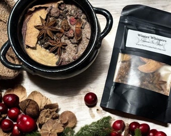 May include: A black ceramic bowl filled with spices and star anise, alongside a black pouch labeled "Winter Whispers". The scene includes scattered cranberries, pine sprigs, and other natural elements, creating a festive, seasonal arrangement.