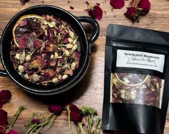 May include: A black pouch with the text "Bewitched Blossoms" and "Sacred Bath Blend" on the front. The pouch contains a blend of dried rose petals and jasmine flowers. The pouch is sitting on a wooden surface with rose petals scattered around it.