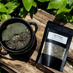 May include: A black cauldron filled with dried herbs and two bay leaves, next to a black pouch labeled "Lunar Dreams" with a visible blend of herbs. The items are arranged on a weathered wooden surface, surrounded by green foliage.