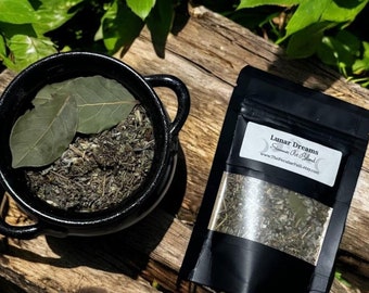 May include: A black cauldron filled with dried herbs and two bay leaves, next to a black pouch labeled "Lunar Dreams" with a visible blend of herbs. The items are arranged on a weathered wooden surface, surrounded by green foliage.