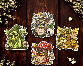 May include: Four whimsical stickers featuring stylized creatures with large eyes and various adornments. One is green with a staff, another has a mushroom cap, a third has a skull, and the last has a flower crown. The stickers are on a dark wood surface.