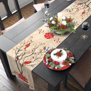 May include: A beige table runner with a winter bird and floral design. The runner features red cardinals, bluebirds, and poinsettias on a beige background. The runner is laid on a dark wood table with a centerpiece of red berries and greenery.