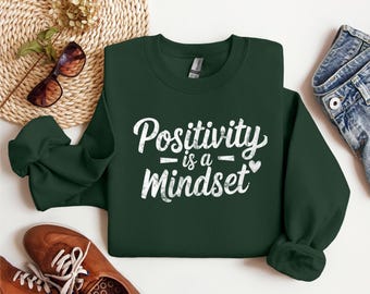 May include: A dark green sweatshirt with the text "Positivity is a Mindset" in white lettering with a distressed look. The text is in a cursive font with a heart symbol after the word "Mindset".