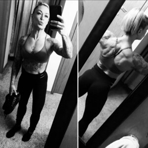 May include: A black and white photo of a person wearing a black sports bra and black pants. The person is standing in front of a mirror and is taking a selfie. The person has tattoos on their arms and back.