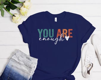 Może przedstawiać: Koszulka w kolorze granatowym z napisem "YOU ARE enough" w kolorowych, vintage'owych literach z małym białym sercem.